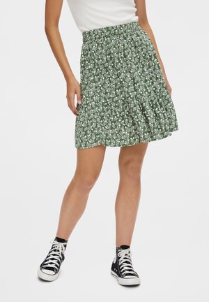 Vrouw draagt een groene bloemen A-lijn rok gecombineerd met zwarte hoge sneakers en een wit mouwloos topje tegen een effen achtergrond.