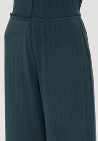 Combinaison côtelée en teal avec une surface texturée, détail de bouton en haut et accent effiloché à la taille, présentant une coupe ample et des jambes larges.