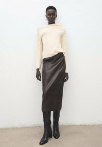 Off-white, kabelstrikket sweater parret med en mørkebrun læder pencilskørt og tætsiddende sorte knæhøje støvler, sat op mod en ensfarvet baggrund.