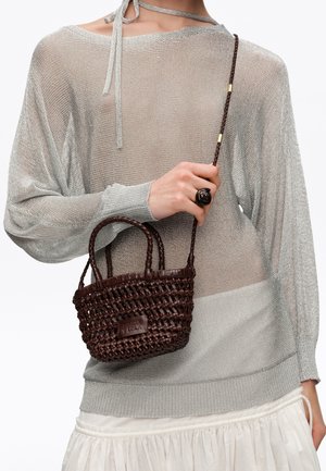 Osoba nosząca przezroczysty szary dziergany top i białą spódnicę, trzymająca małą ciemnobrązową plecioną torebkę z podwójnymi uchwytami i długim plecionym paskiem.