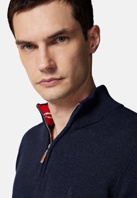 Granatowy sweter z dzianiny z wysokim kołnierzem, z czerwono-białym paskowanym wnętrzem, pół-zamkiem oraz subtelnym detalem z logo.