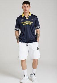 Tricou sportiv de marine cu guler galben și text "FOOTBALL LEAGUE", asortat cu pantaloni scurți albi ce prezintă un logo. Include accente de manșetă cu model.