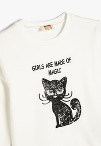 Felpa bianca con una grafica di un gatto nera in paillettes con baffi, e il testo "LE RAGAZZE SONO FATTE DI MAGIA" in lettere nere.