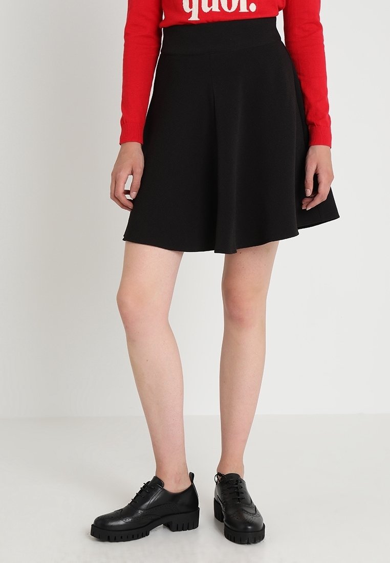 Femme portant une jupe noire évasée, des chaussures noires épaisses à lacets et un haut à manches longues rouge, debout devant un fond blanc.