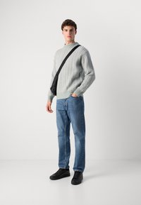 Young man wearing light gray cable knit sweater, blue jeans, black sneakers, and a black crossbody bag, standing against a plain white background.