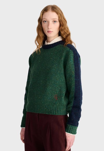 Jeune femme aux cheveux roux ondulés portant un pull en maille vert avec des manches bleu marine et un pantalon rouge foncé, debout devant un fond uni.