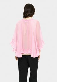Light pink sheer cape with ruffled edges, gathered neckline, and loose, flowing design; worn over a lighter base layer and dark pants.