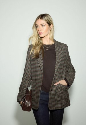 Karo-Blazer in Brauntönen und Grün, sichtbare Stoffstruktur, kombiniert mit einem dunklen Pullover und Jeans, hält eine braune Handtasche.
