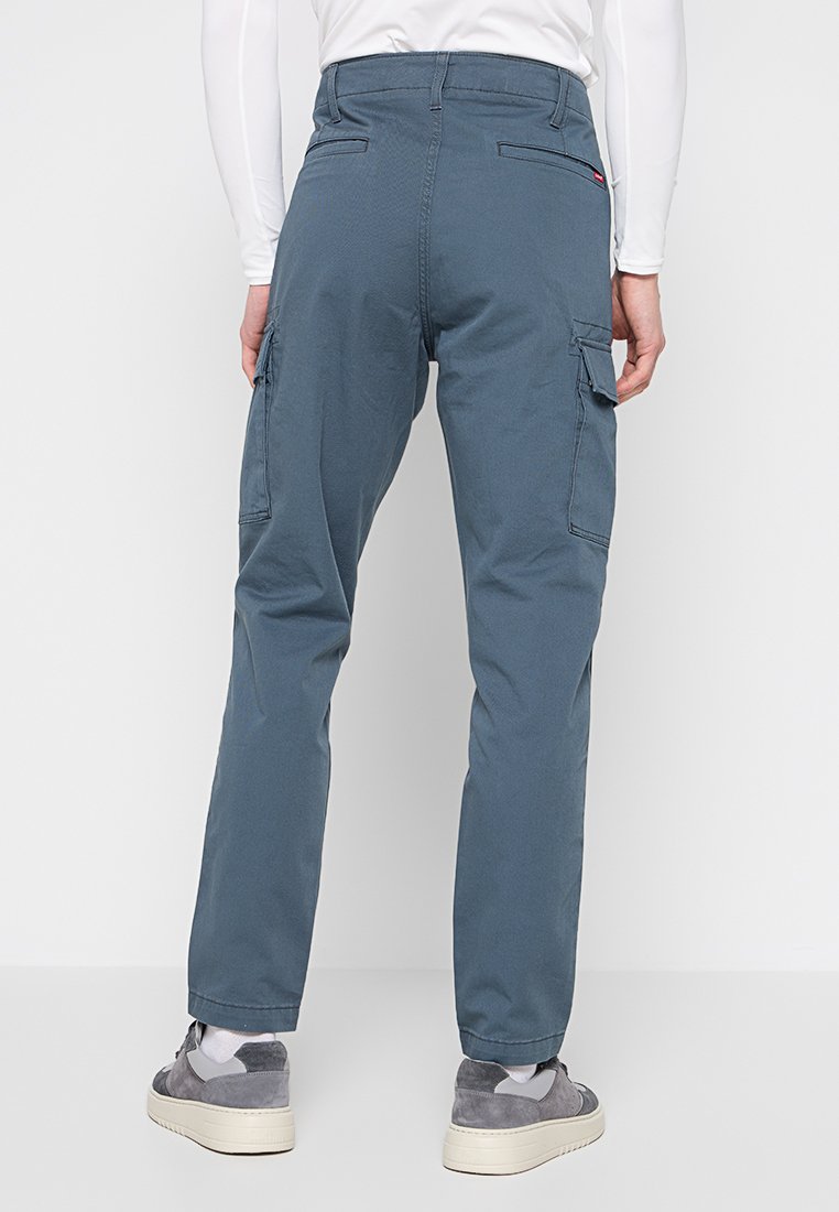 Cargo-Hosen aus dunkelblauem Stoff mit zwei Seitentaschen, Nahtakzenten und einer lässigen Passform. Kombiniert mit grauen Sneakers.