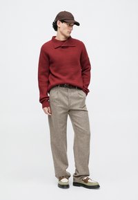 Bărbat îmbrăcat cu un pulover tricotat roșu, pantaloni bej cu o curea maro, încălțat cu adidași verzi și albi, purtând o șapcă de baseball maro, stând și uitându-se în lateral.