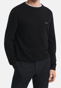 Pull noir texturé avec un col rond et des poignets côtelés. Présente un col intérieur gris et un petit logo blanc sur la poitrine. Associé à un pantalon foncé.