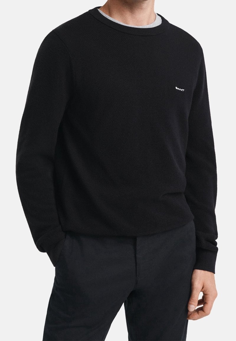 Pull noir texturé avec un col rond et des poignets côtelés. Présente un col intérieur gris et un petit logo blanc sur la poitrine. Associé à un pantalon foncé.