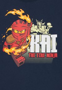 Marinblå T-shirt med en grafik av en röd ninja med lågor, märkt "KAI THE FIRE NINJA," på en strukturerad bakgrund med ljusa accenter.