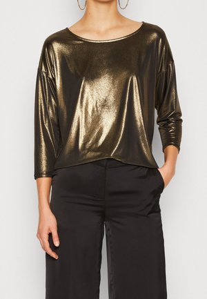 Kobieta w błyszczącej, metalicznej złotej bluzce z długim rękawem i czarnych szerokich spodniach, z jedną ręką w kieszeni i widocznymi kolczykami kołami.