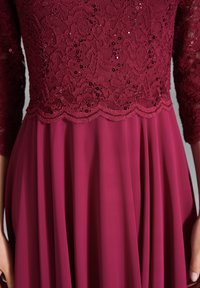 Robe bordeaux avec un corsage en dentelle ornée de sequins, bords en vagues, et une jupe plissée fluide. Manches longues en dentelle.