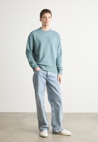 Sudadera azul claro con cuello redondo, de corte relajado, combinada con unos jeans de mezclilla claros y holgados y zapatillas blancas. Textura suave.