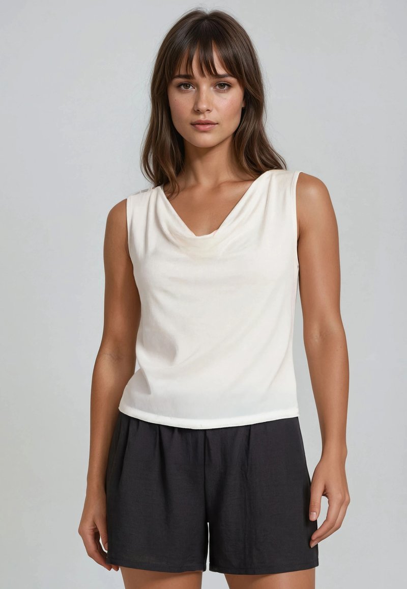 Femme aux cheveux bruns portant un haut blanc sans manches et un short noir ample, debout devant un fond gris clair uni.