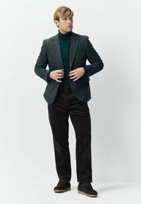 Man wearing dark green turtleneck, grey blazer, dark brown corduroy pants, and black shoes, adjusting blazer buttons while looking sideways.