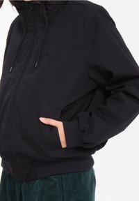 Giacca nera con zip caratterizzata da un colletto alto, polsini e orlo a costine, tasche laterali e un logo minimalista sul petto. Tessuto dalla texture liscia.