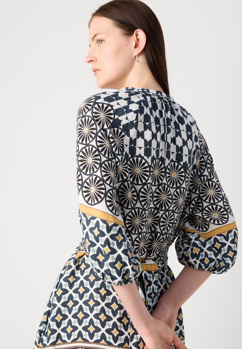 Bunt gemustertes Kleid mit geometrischen Designs in Schwarz, Weiß und Gelb. Es hat einen Bindegürtel in der Taille und dreiviertellange Ärmel.