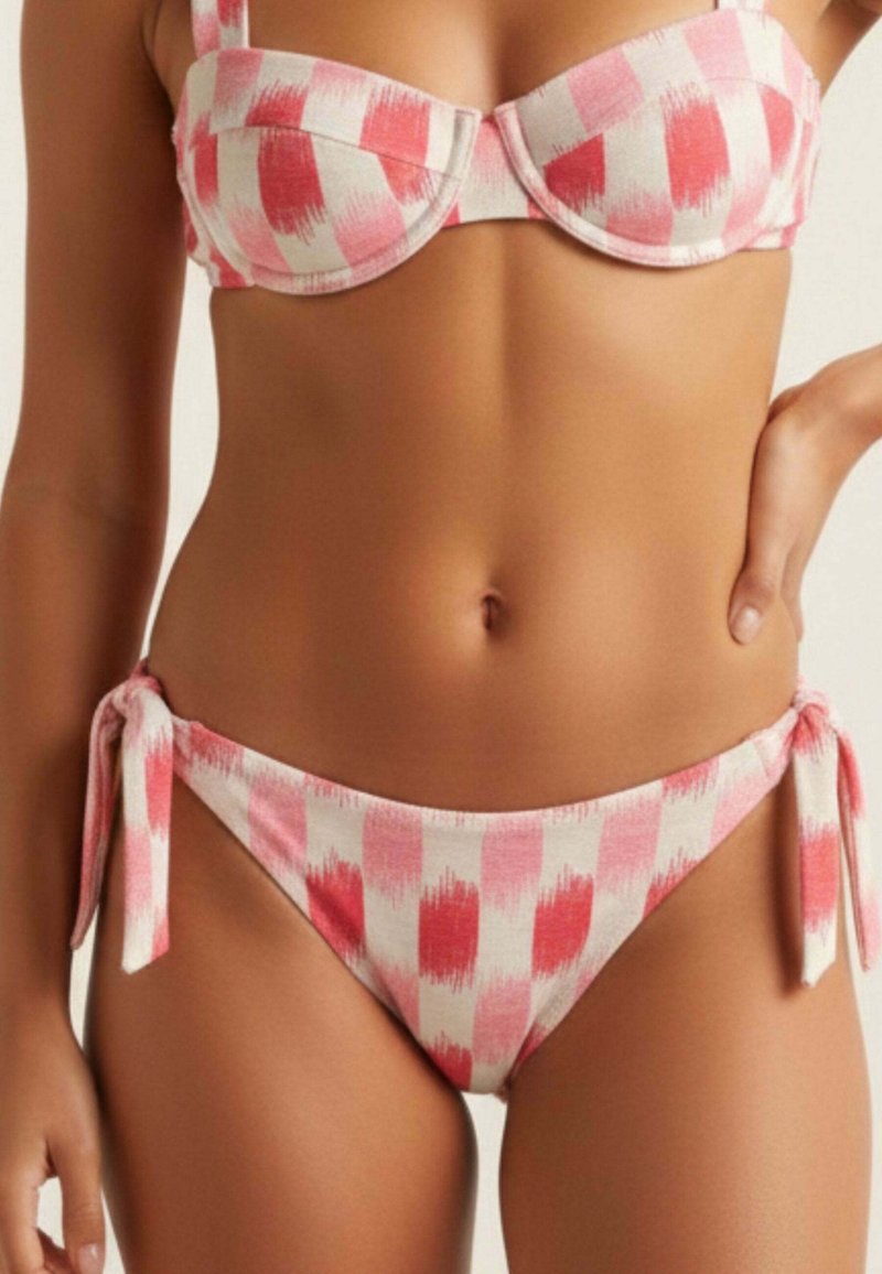 Mujer con bikini a cuadros rosa y blanco con lazos laterales, mostrando el torso y parte de los brazos contra un fondo neutro.