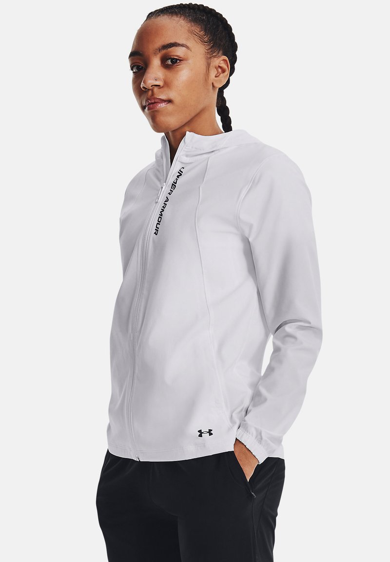 Witte sportjas met ritssluiting aan de voorkant, hoge kraag en zwarte logo-accenten. Glad, lichtgewicht materiaal met een aansluitend ontwerp, lange mouwen en zakken.