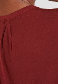 Burgundowa bluzka o gładkiej teksturze, zaokrąglonym dekolcie i subtelnym detalem plisowania z tyłu. Materiał wydaje się lekki i dopasowany.