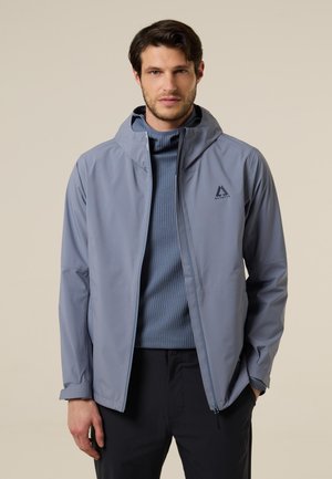 Hombre vistiendo una chaqueta con capucha gris claro sobre un suéter azul texturizado y pantalones negros, de pie frente a un fondo beige liso.