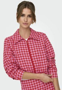 Roze en rode houndstooth ritsjack met een grote kraag en lange mouwen, met een textuurpatroon en contrasterende ritsdetails.
