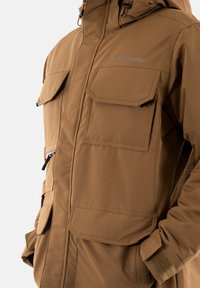Chaqueta impermeable marrón con múltiples bolsillos frontales, un bolsillo en el pecho con cremallera, capucha ajustable y puños de velcro. Textura suave, tela duradera.