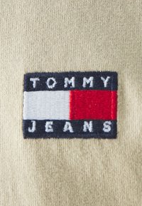 Tommy Jeans hímzett címke, amely tengerészkék, fehér és piros színblokkokból áll, a "TOMMY" és "JEANS" felirattal egy bézs, textúrált anyagon.