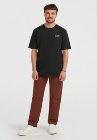 Sort kortærmet t-shirt med et lille hvidt logo på venstre bryst, sammen med rustfarvede bukser og hvide sneakers.