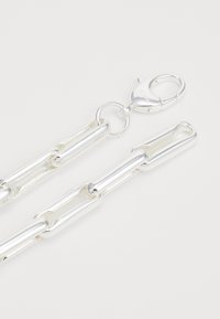 Collier en chaîne en métal argenté avec des maillons rectangulaires allongés et un fermoir en mousqueton sur fond blanc.
