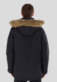Man in een zwarte winterjas met een met bont afgewerkte capuchon, gezien vanaf de achterkant tegen een effen achtergrond.