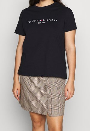 Osoba ubrana w czarną koszulkę Tommy Hilfiger oraz beżową, kratowaną spódnicę z wysokim stanem, stojąca na jednolitym tle.