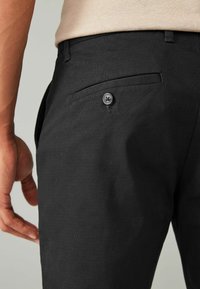 Pantalon noir texturé avec une poche arrière fermée par un bouton. Le tissu semble durable avec un motif tissé subtil.