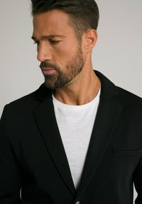 Blazer nero con rever a punta, chiusura a un bottone e taschino sul petto, sovrapposto a una maglietta bianca con una sottile trama.