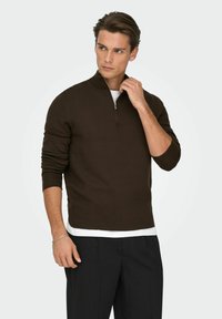 Pull marron avec col haut et fermeture éclair partielle, en matière tricotée douce, porté sur une chemise blanche, accompagné d'un pantalon noir.