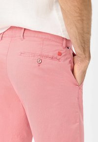 Pantaloni rosa con texture, dotati di una tasca posteriore, chiusura con bottoni e un piccolo etichetta del marchio sulla vita. Vestibilità su misura con tasche laterali.