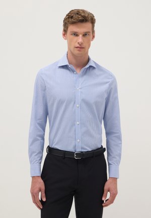 Homme portant une chemise à rayures bleu clair boutonnée, une ceinture noire et un pantalon noir, debout devant un fond uni.