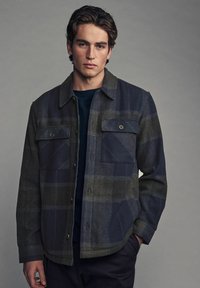Flanelová overshirt v námorníckej a zelenej kárované vzore. Obsahuje dve náprsné vrecká a zapínanie na gombíky. Textúrovaná tkanina a klasický dizajn goliera.
