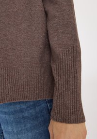 Brauner Strickpullover mit gerippten Bündchen, der eine weiche Textur und einen lässigen Sitz zeigt. Der Ärmel reicht leicht über das Handgelenk hinaus.