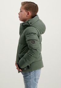 Groene geïsoleerde pufferjas met een kap, voorzien van een logopatch op de linkerarm en zijzakken, gecombineerd met lichtblauwe spijkerbroek.