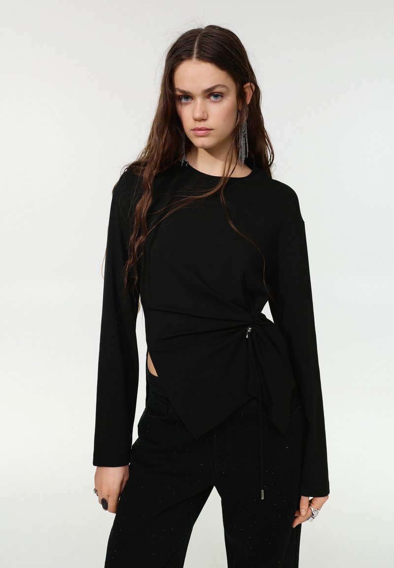 Haut noir à manches longues avec un design torsadé, ouverture latérale et détail à cordon de serrage à la taille. Présente une texture lisse et une coupe décontractée.