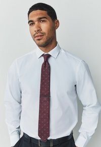 Weißes Hemd mit spitzem Kragen, kombiniert mit einer bordeauxfarbenen Krawatte mit Rauten-Muster und Clip. Ein schwarzer Gürtel betont das Outfit.