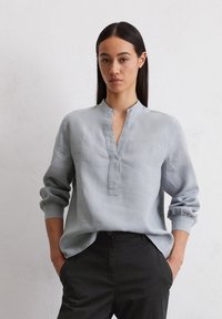 Chemise en lin bleu clair avec col mandarin, patte de boutonnage sur le devant et manches froncées. Associée à un pantalon noir, avec une coupe décontractée.