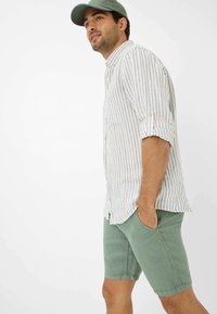 Camicia a righe di colore chiaro con maniche arrotolate abbinata a pantaloni corti verdi. L'outfit presenta una vestibilità casual e un design minimalista.
