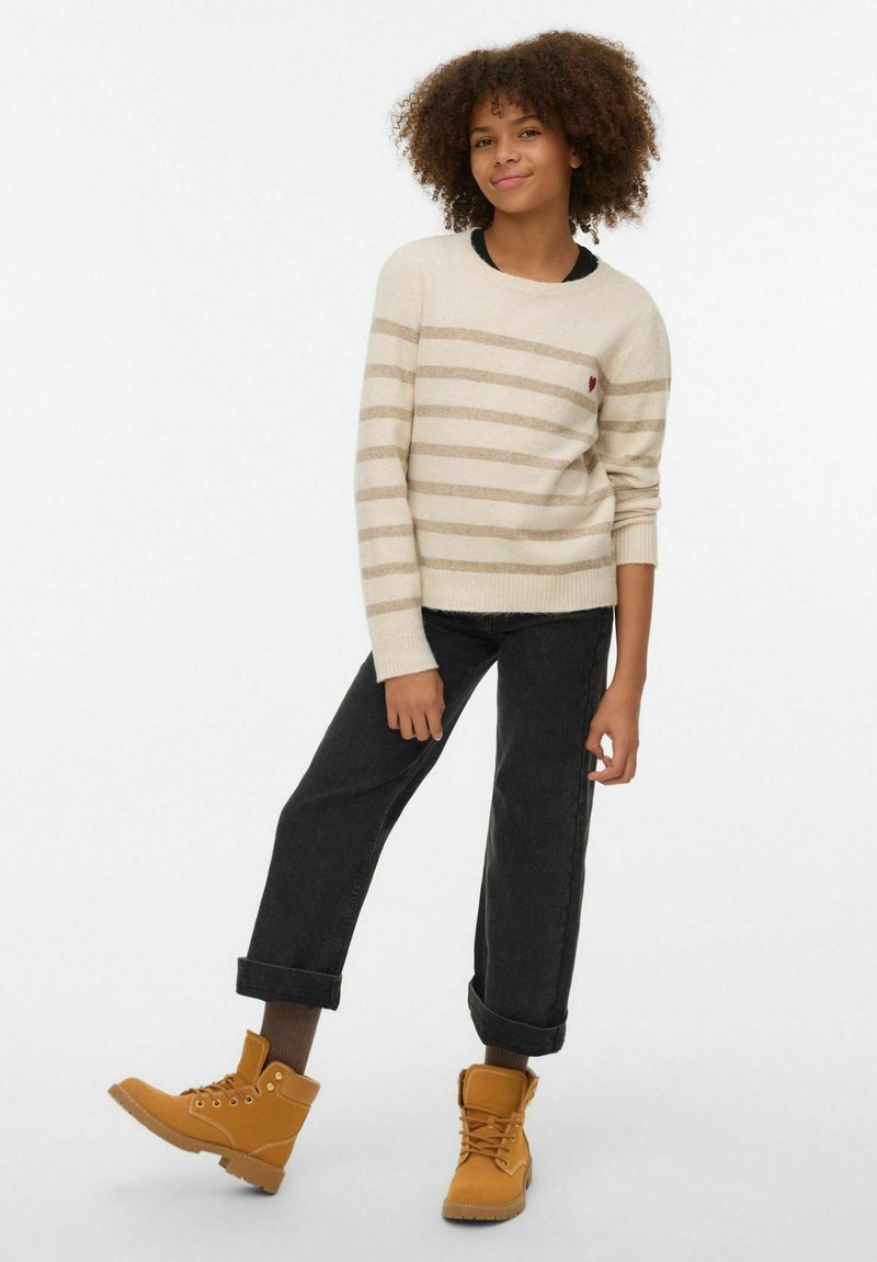 Pull à rayures beige avec un col rond, associé à un jean noir et des bottines jaunes. Le pull comporte un petit logo en forme de cœur.