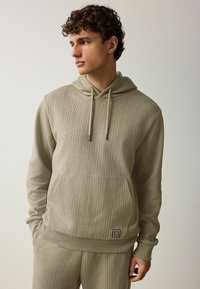 Sudadera con capucha de manga larga en verde olivo claro, que cuenta con tejido texturizado, un bolsillo frontal y una pequeña etiqueta de logo en el dobladillo.