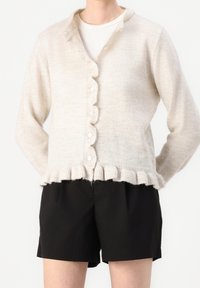 Cárdigan de punto beige con un dobladillo de pico, que presenta un cierre frontal de botones, puños acanalados y una textura suave, llevado sobre una blusa blanca y pantalones cortos negros.